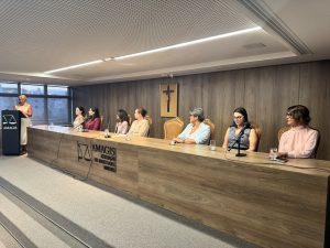 A imagem mostra uma mesa de honra composta por sete mulheres no auditório da Amagis. As mulheres estão sentadas atrás de uma mesa de madeira com microfones. À frente da bancada está a logomarca da Amagis. A segunda mulher da direita para a esquerda é a juíza Carolina Aleixo Benétti de Oliveira Rodrigues. À esquerda, uma mulher está em pé atrás de uma tribuna, que também exibe a logomarca da Amagis. Ao fundo, há uma parede de madeira com um crucifixo ao centro.
