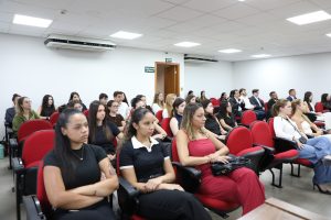 A imagem mostra os alunos da Faculdade de Direito Milton Campos durante sessão no Tribunal Pleno. Eles estão sentados em cadeiras vermelhas do Plenário do TJMMG. As paredes ao fundo são brancas e há uma bancada madeira em primeiro plano
