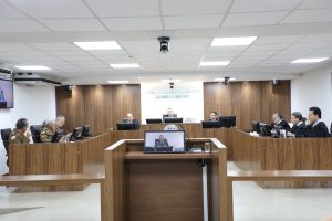 A imagem mostra os desembargadores durante a Sessão de Julgamento no Tribunal Pleno, sentados em uma tribuna de madeira. Eles estão posicionados atrás de microfones e computadores, alguns com trajes militares, outros de toga e um de camisa social e gravata. O ambiente apresenta paredes revestidas em madeira, bandeiras oficiais e o brasão do TJMMG ao fundo.
