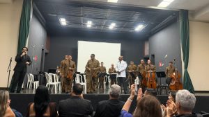 A imagem mostra a OSPM no palco, com músicos em pé vestindo fardas bege da PMMG e segurando instrumentos como violinos e violoncelos. O maestro está ao centro, de farda branca. Também sobre o palco, à esquerda da imagem, um homem vestindo terno preto discursa. O público está em primeiro plano na imagem, de frente ao palco, todos aplaudindo de pé.