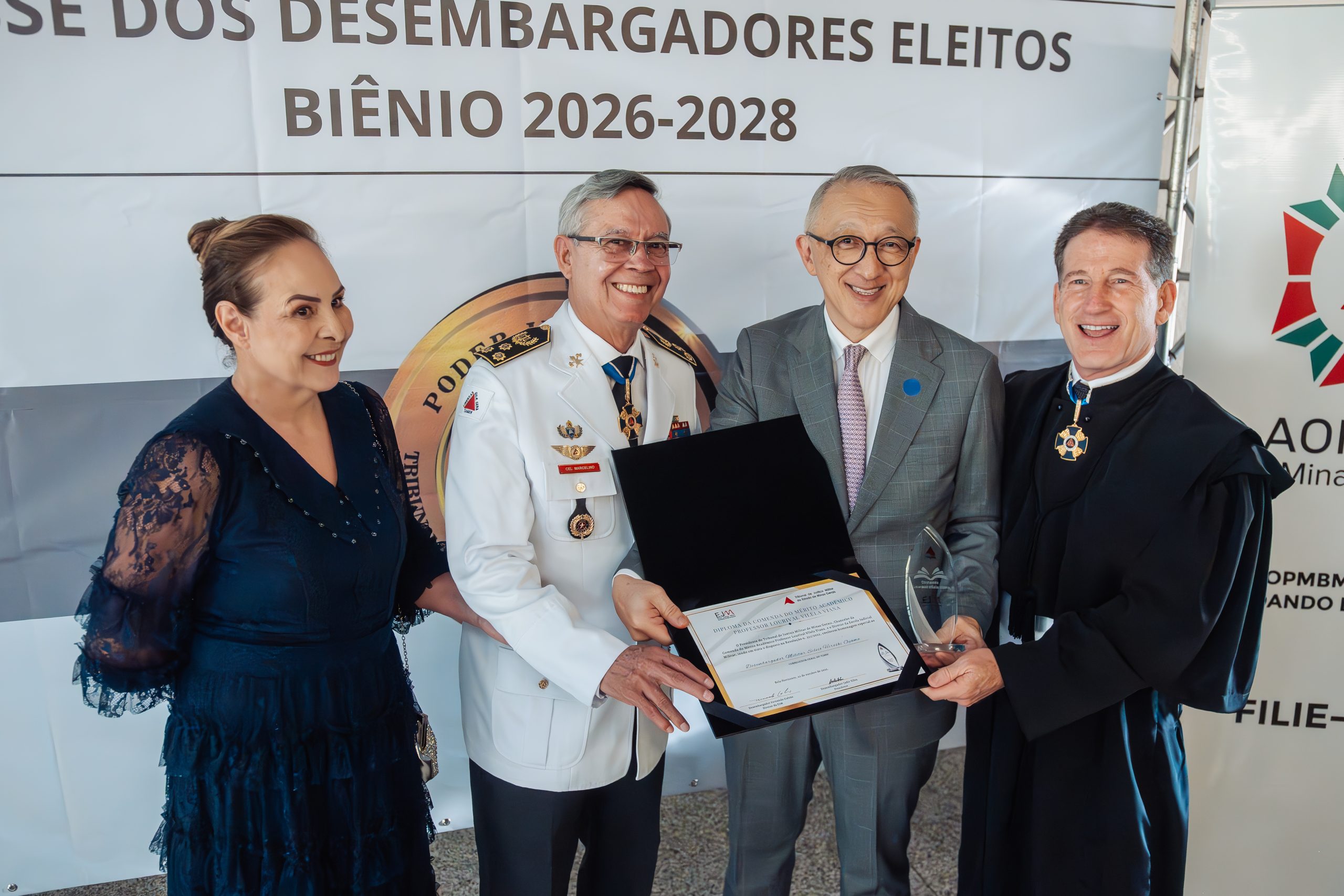 A imagem mostra quatro pessoas em pé. Da direita para a esquerda, estão o desembargador Fernando Antônio Nogueira Galvão da Rocha, vestindo toga preta, segurando uma comenda junto ao presidente do TJMSP, desembargador Silvio Hiroshi Oyama, que usa terno cinza, e também segura um diploma ao lado do presidente do TJMMG, desembargador Osmar Duarte Marcelino, que está trajando uniforme branco do Corpo de Bombeiros Militar de Minas Gerais. Ao lado dele está a esposa, Olga de Lourdes Aquino Duarte, usando vestido azul e tem os cabelos presos em um coque alto. Ao fundo, há um painel escrito “Posse dos desembargadores eleitos biênio dois mil e vinte e seis dois mil e vinte e oito