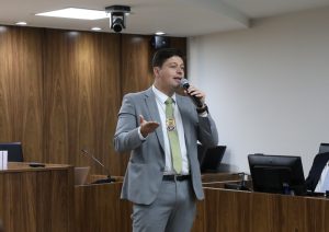 A imagem mostra Bruno de Almeida Felipe em pé, na sala do Pleno do TJMMG, vestindo terno cinza, camisa branca e gravata verde, segurando um microfone. Ele usa um distintivo pendurado no pescoço. Ao fundo há um painel e uma tribuna de madeira
