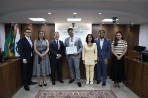 A imagem mostra sete pessoas em pé, entre homens e mulheres, no Plenário do TJMMG. Da esquerda para a direita, estão: o juiz Bruno Cortez Torres Castelo Branco; a juíza Carolina Aleixo Benetti de Oliveira Rodrigues; o desembargador Fernando Antônio Nogueira Galvão da Rocha; Bruno de Almeida Felipe; a juíza Renata Rodrigues de Pádua; o juiz João Pedro Hoffert Monteiro de Lima; e a coordenadora da EJM, Ana Paula Araújo Sales Rezende. Ao fundo, na parede, está escrito “Tribunal de Justiça Militar de Minas Gerais” e, à esquerda, aparecem bandeiras.