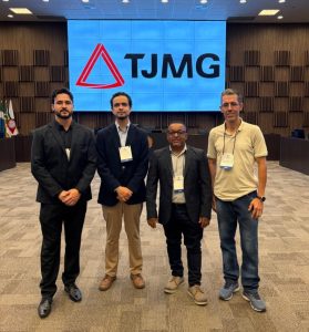 A imagem mostra a comitiva do TJMMG em pé, ao centro do auditório do TJMG, tendo ao fundo paredes de madeira e um telão com a marca do tribunal. Da esquerda para a direita estão o servidor Thiago Augusto Duarte Pereira, de terno preto; o juiz de Direito George Walter Barreto Paviotti, de blazer azul escuro e calça bege; o servidor Edivaldo Pereira dos Santos, de terno escuro e camisa social branca, e Altino Couto Machado, de camisa amarela e calça jeans.