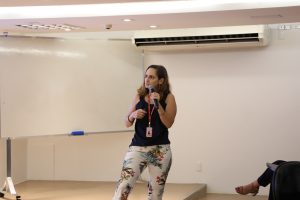 A imagem mostra a servidora Ana Paula Hermont em pé no palco do auditório do TJMMG, durante o encerramento do evento. Ela veste blusa preta, calça branca com estampa florida e segura um microfone. Ela está posicionada à frente do espaço de apresentação.