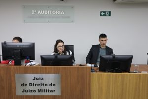 A imagem mostra a juíza de Direito Carolina Aleixo Benétti de Oliveira Rodrigues ao centro de uma bancada de madeira durante abertura dos trabalhos de Autoinspeção da auditoria. A juíza veste roupa com estampa branca e preta. À direita da imagem, de terno preto e camisa cinza, está o juiz de Direito substituto Antônio Moreno Boregas e Rêgo. Na frente da bancada de madeira está uma placa escrita juiz de direito do juízo militar, e ao fundo, na parede, uma placa de acrílico com o nome da auditoria