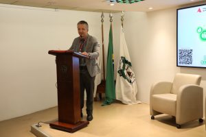 A imagem mostra o secretário especial da Presidência, Giovâni Viana Mendes, em pé atrás de uma tribuna de madeira, falando ao microfone no auditório do TJMMG. Ele veste blêiser cinza claro e calça cinza escuro. Ao fundo, aparecem três bandeiras. À direita da imagem há uma poltrona clara e parte de uma tela de apresentação fixada na parede.