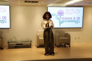 A imagem mostra Edilene Nascimento, em pé, no centro do auditório do TJMMG. Ela veste blusa branca e calça marrom. Ao fundo, aparecem duas telas de apresentação fixadas na parede.
