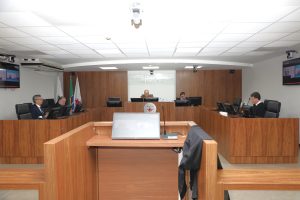 A imagem mostra os desembargadores durante a Sessão de Julgamento da primeira Câmara, sentados em uma tribuna de madeira. Eles estão posicionados atrás de microfones e computadores. Ao centro está o desembargador Rúbio Paulino Coelho, usando uniforme militar, e o diretor judiciário Eli Alvarenga, usando roupa preta. À esquerda estão os desembargadores Osmar Duarte Marcelino, vestindo terno azul escuro, e Jadir Silva, vestindo toga preta. À direita está o desembargador Fernando Galvão, usando toga preta. O ambiente apresenta paredes revestidas em madeira, bandeiras oficiais e o brasão do TJMMG ao fundo. 