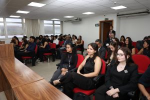 A imagem mostra os alunos da Faculdade de Direito da UFMG durante sessão de julgamento da primeira Câmara. Eles estão sentados em cadeiras vermelhas do Plenário do TJMMG. As paredes ao fundo são brancas, com amplas janelas à esquerda da imagem, e há uma bancada madeira em primeiro plano.