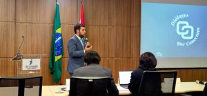 A imagem mostra o juiz de Direito João Pedro Hoffert Monteiro de Lima vestindo um terno azul, camisa branca e gravata cinza. O juiz está falando para uma plateia que aparece de costas na imagem, e ao fundo há uma parede de madeira com um telão azul exibindo o nome do evento.