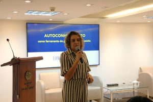 A imagem mostra a tenente-coronel PM psicóloga Eliene Lima, em pé, falando ao microfone no auditório do TJMMG. Ele veste blusa e calça listradas. Ao fundo, aparece uma tela de apresentação fixada na parede, com o nome da palestra. À esquerda da imagem há uma tribuna de madeira.
