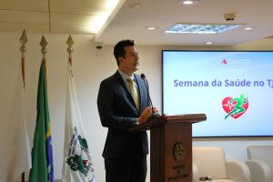 A imagem mostra o desembargador Fernando José Armando Ribeiro, em pé atrás de uma tribuna de madeira, falando ao microfone no auditório do TJMMG. Ele veste terno escuro e gravata amarela. Ao fundo, aparecem três bandeiras. À direita da imagem há duas poltronas claras e parte de uma tela de apresentação fixada na parede, com identidade visual do evento.