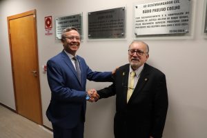 A imagem mostra o desembargador Osmar Duarte Marcelino, vestindo terno azul, cumprimentando com um aperto de mão o desembargador Jadir Silva, que está de terno preto. Ambos estão em pé diante de uma parede com placas de ex-presidentes, no Salão Nobre do TJMMG.
