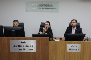 A imagem mostra a Juíza de Direito Daniela de Freitas Marques ao centro de uma bancada de madeira durante abertura dos trabalhos de Autoinspeção da auditoria. A juíza veste uma toga, usa óculos e fala ao microfone. À direita da imagem está o juiz de Direito João Pedro Hoffert Monteiro de Lima, de terno escuro, camisa azul clara e gravata vermelha clara. Do lado esquerdo da imagem está o gerente de Secretaria Marcos Roberto Maciel, de camisa preta. Atrás deles está uma placa de acrílico com o nome da 3ª Auditoria