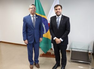 A imagem mostra o presidente do TJMMG, desembargador Osmar Duarte Marcelino, vestindo terno azul, ao lado do juiz de Direito do Juízo Militar João Pedro Hoffert Monteiro de Lima, que usa terno preto, na sala da Presidência do TJMMG. Ao fundo, há duas bandeiras.