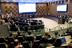 A imagem mostra o auditório do Tribunal Pleno do TJMG. Um homem fala ao microfone, cercado pela plateia sentada em cadeiras assistindo a apresentação. O ambiente tem paredes de madeira.