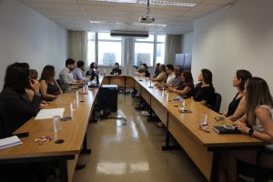 Imagem mostra uma sala de reunião com pessoas, entre homens e mulheres, sentadas diante de uma mesa de madeira em formato de “U”. Sobre a mesa, há copos de água, papéis e canetas. A sala tem paredes brancas, com uma ampla janela ao fundo.