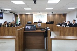 A imagem mostra os desembargadores durante a Sessão de Julgamento no Tribunal Pleno, sentados em uma tribuna de madeira. Eles estão posicionados atrás de microfones e computadores, alguns com trajes militares, outros de toga. O ambiente apresenta paredes revestidas em madeira, bandeiras oficiais e o brasão do TJMMG ao fundo.