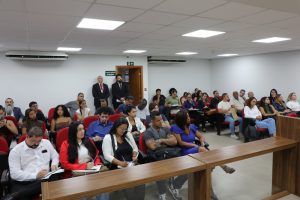 A imagem mostra os alunos da Faculdade de Direito Estácio de Sá durante sessão no Tribunal Pleno. Eles estão sentados em cadeiras vermelhas do Plenário do TJMMG. As paredes ao fundo são brancas e há uma bancada madeira em primeiro plano.