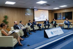A imagem mostra o palco do auditório do Conselho Nacional de Justiça, com sete homens e uma mulher reunidos em poltronas claras, todos vestindo trajes formais. Da direita para a esquerda, está o juiz de Direito do Juízo Militar, João Pedro Hoffert Monteiro de Lima, vestindo terno azul e falando ao microfone. Ao fundo, as paredes são revestidas de madeira. Há um telão exibindo a imagem do magistrado e, à esquerda, aparece a logomarca do CNJ.