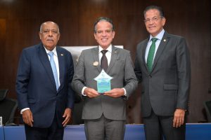 A imagem mostra três homens em pé em um palco. O primeiro à esquerda é o ministro Benedito Gonçalves, que veste terno e gravata azul, e camisa branca. Ao centro está o desembargador Jâmes Ferreira Santos. Ele veste um terno cinza com gravata preta e segura o troféu da Corregedoria Ética nas mãos. O troféu é de acrílico, com tons de azul. À direita está o ministro Mauro Campbell Marques, de terno cinza, camisa azul e gravata verde.