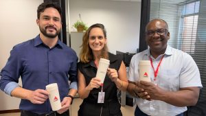 A imagem mostra três servidores do TJMMG, sendo dois homens e uma mulher ao centro, segurando os cópos personalizados sustentáveis. Eles estão em um ambiente interno de uma sala no Tribunal. Os cópos são em tons claros e de um lado exibem a logomarca do TJMMG; do outro, o nome de cada servidor