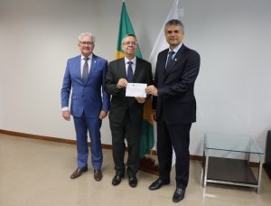 A imagem mostra três homens em pé, um ao lado do outro, na sala da Presidência do TJMMG, tendo ao fundo duas bandeiras. Ao centro está o desembargador Osmar Duarte Marcelino, vestindo terno preto e gravata azul. Ele segura o convite da posse dos desembargadores do TRE-MG. À direita da imagem está o desembargador Carlos Henrique Perpétuo Braga, vestindo terno e gravata preta. Ele também segura o convite da posse. Já à esquerda da imagem, está o desembargador Sálvio Chaves, vestindo terno azul e gravata cinza escuro.