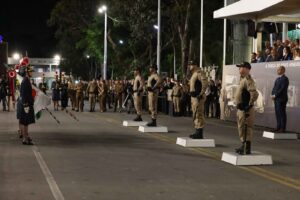 A imagem mostra a solenidade de transmissão dos cargos da PMMG, no pátio da Academia de Polícia Militar. Em primeiro plano, quatro policiais militares uniformizados estão posicionados sobre pequenos palanques brancos, em posição de continência. Atrás deles, aparece o governador de Minas Gerais, também em um pequeno palanque. À esquerda da imagem, estão duas pessoas com uniformes cerimoniais militares segurando bandeiras, voltadas para os policiais. Ao fundo, há uma plateia com o público observando o evento, além de autoridades em um palco coberto à direita.