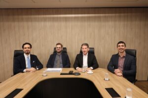 A imagem mostra quatro homens sentados na sala Diamante do TJMMG, dispostos em uma mesa de reunião em tons de madeira e preto, mesmas cores do painel ao fundo. Da direita para a esquerda está o juiz Antônio Moreno Boregas e Rego, vestindo terno preto e camisa marrom claro; o desembargador Fernando Antônio Nogueira Galvão da Rocha, vestindo terno preto e camisa branca; o professor André Pedrolli Serretti, usando terno preto e camisa cinza, de óculos; e o juiz George Walter Barreto Paviotti, usando também um terno preto e camisa branca com gravata azul.