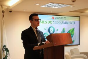 A imagem mostra o juiz de Direito do Juízo Militar Bruno Cortez Torres Castelo Branco, em pé atrás de uma tribuna de madeira, falando ao microfone no auditório do TJMMG. Ele veste um terno preto, camisa clara e gravata escura. Ao fundo, aparece uma tela de apresentação fixada na parede, com a imagem do cartaz do evento.