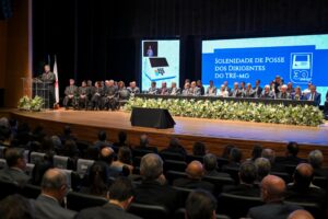 A imagem mostra o auditório do Teatro do Minas Tênis Clube durante a cerimônia de posse dos novos dirigentes do TRE-MG. No palco, estão diversas autoridades sentadas em uma longa mesa decorada com arranjos de flores brancas. À esquerda, uma pessoa fala em uma tribuna transparente, com bandeiras posicionadas atrás. Ao fundo, estão dois grandes telões, sendo o da direita com a imagem da urna eletrônica e, no painel central, escrito “Solenidade de Posse dos Dirigentes do TRE-MG” 