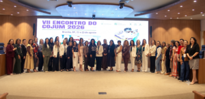 A imagem mostra um grupo de mulheres, posicionadas uma ao lado da outra em um auditório do TJES. Todas vestem trajes sociais em diferentes cores e estilos. A terceira pessoa da direita para a esquerda da imagem é a juíza de Direito do Juízo Militar Daniela de Freitas Marques, usando blusa vermelha. Ao fundo, há um grande telão escrito: “sétimo encontro do cojum 2026”. O auditório possui paredes revestidas de madeira, teto com iluminação embutida e um piso claro
