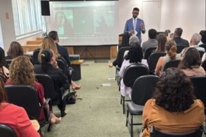 A imagem mostra uma sala institucional, com várias pessoas sentadas em cadeiras pretas, voltadas para o conselheiro Fábio Francisco Esteves, que está em pé à frente da plateia. Ele veste terno azul e fala ao público enquanto gesticula. Ao fundo, aparece uma tela de projeção exibindo uma videoconferência com participantes on-line.