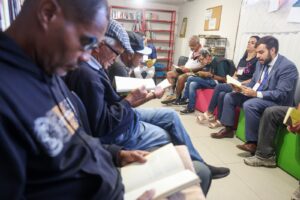 A imagem mostra um grupo de cerca de nove pessoas reunidas em no clube de leitura do Inaper. Elas estão sentada em uma fileira de cadeiras e pufes coloridos, todas concentradas na leitura de livros. O ambiente tem piso claro, paredes brancas e, ao fundo, destacam-se estantes de metal vermelho repletas de livros. O primeiro à direita da imagem é o juiz de Direito do Juízo Militar João Pedro Hoffert Monteiro de Lima, vestido com um terno azul-escuro, camisa social branca, gravata azul-escura e sapatos sociais de couro marrom. O juiz segura um livro aberto e faz a leitura para o grupo. Em primeiro plano, à esquerda, a lateral do corpo e o rosto de um homem negro vestindo um casaco de moletom preto aparecem em corte fechado e levemente desfocados, também com os olhos voltados para um livro aberto em suas mãos. Os demais participantes, de faixa etária diversa — incluindo senhores mais velhos de boné e boina —, completam a roda de leitura, criando uma atmosfera de atenção compartilhada.
