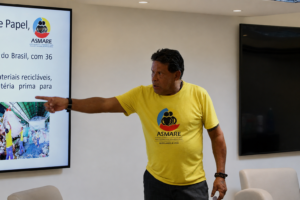 A imagem mostra o palestrante Alfredo de Sousa Matos em pé, no auditório do TJMMG. Ele veste uma blusa amarela e calça preta. Com o braço esquerdo estendido, ele aponta para uma tela de apresentação posicionada à esquerda da imagem projetando dados fornecidos pela Asmare.
