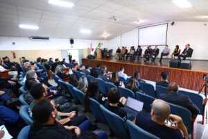 A imagem mostra um auditório na Academia de Bombeiros Militar de Minas Gerais. Em primeiro plano, o público aparece sentado em fileiras de cadeiras na cor azul. Ao fundo, aparece um palco com piso de madeira escura, com doze autoridades sentadas em cadeiras, entre homens e mulheres. Entre as autoridades,está o juiz de Direito titular do Juízo Militar, Marcos Luiz Nery Filho. À esquerda, um homem discursa em pé atrás de uma tribuna de madeira. A parede ao fundo é branca e possui uma grande tela de projeção fixada.