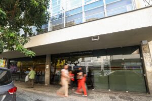 A imagem mostra a fachada do Ciapop-BH, um edifício com portas e janelas de vidro na fachada, e uma ampla marquise de concreto. Logo acima da entrada de vidro, está escrito "Poder Judiciário do Estado de Minas Gerais". Na calçada estão pedestres caminhando com a imagem borrada, sugerindo movimento. 