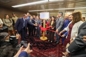 A imagem mostra um grupo de pessoas, entre homens e mulheres, reunidas em um ambiente interno no Ciapop-BH. Todos vestem trajes formais. No centro, há uma placa de inauguração na cor branca, fixada em um pedestal de madeira. As pessoas estão posicionadas em semicírculo ao redor da placa, e descerram o dispositivo coberto com um tecido vermelho. Entre as autoridades presentes, a segunda pessoa à direita da imagem é o juiz de Direito João Pedro Hoffert Monteiro de Lima, de terno azul. O local possui um tapete vermelho e o teto com luminárias embutidas.  