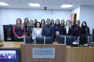 A imagem mostra um grupo de 13 mulheres e um homem que integram a 4ª AJME. Todos estão em pé, atrás de uma bancada de madeira. Na parede atrás deles está uma placa de acrílico escrito: Quarta auditoria. Justiça Militar de Minas Gerais. O homem é o juiz André de Mourão Motta, que veste terno escuro, camisa preta e gravata listrada em preto e marrom. Na imagem, do lado esquerdo do juiz está a juíza Renata Rodrigues de Pádua, de roupa cinza claro. As demais mulheres da imagem, trajadas de maneira formal, são servidoras e estagiárias da auditoria. 