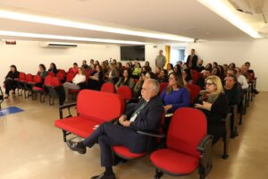 A imagem mostra o auditório do TJMMG com uma plateia de homens e mulheres sentados em fileiras de poltronas vermelhas. Na primeira fileira, em destaque, está o desembargador Rúbio Paulino Coelho, que usa um terno azul-marinho, camisa social branca, gravata verde-escura e sapato social marrom. O auditório tem tema e paredes brancas.