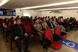 A imagem mostra o auditório do TJMMG com uma plateia de homens e mulheres sentados em fileiras de poltronas vermelhas. Na primeira fileira, sentado na primeira poltrona à direita, está o desembargador Rúbio Paulino Coelho, que usa um terno cinza, camisa social branca, gravata preta e sapato social preto. O auditório tem teto e paredes brancas.