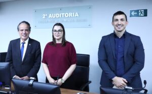 A imagem mostra o desembargador Jâmes Ferreira Santos, vestindo terno preto, camisa branca e gravata azul, a juíza Carolina Aleixo Benétti de Oliveira Rodrigues, vestido blusa na cor vinho e calça preta; e o juiz Antônio Moreno Boregas e Rêgo, vestindo terno e camisa azul escuro. Todos estão em pé, com as mãos cruzadas na frente do corpo, posicionados na sala de audiências da segunda auditoria, um local de parede branca com uma placa de acrílico transparente com o nome da auditoria.