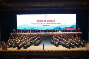 A imagem mostra o interior do Grande Teatro do Palácio das Artes durante a cerimônia de posse da nova direção do TJMG. No palco, diversas autoridades, vestidas com trajes formais, estão alinhadas atrás de uma longa mesa decorada com arranjos de flores. Ao fundo, aparece um grande telão com a identidade visual do evento, escrito “posse da direção – tribunal de justiça do estado de minas gerais – biênio 2026-2028". Nas laterais do palco, autoridades vestidas com togas estão sentadas em cadeiras enfileiradas na diagonal.