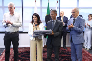 A imagem mostra quatro pessoas em pé no TJMG. Da direita para a esquerda estão o desembargador Rúbio Paulino Coelho, vestindo terno azul e gravata preta, o desembargador Vicente de Oliveira Silva, vestindo terno escuro e gravata cinza, e segurando um cobertor juntamente com a desembargadora Maria Luíza de Marilac, e o assessor da diretoria-geral do TRT-MG, Gustavo Henrique Mendes, vestindo camisa clara e calça preta. Ao fundo, aparecem bandeiras, além de autoridades, entre homens e mulheres.