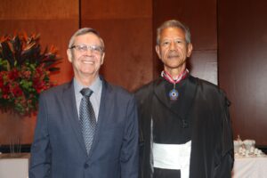 A imagem mostra dois homens posicionados lado a lado do busto para cima, posando para uma foto. Eles estão centralizados contra um fundo composto por painéis de madeira em tons quentes de marrom. O primeiro à esquerda da imagem é o presidente do TJMMG, que usa óculos de grau com armação retangular escura. Está vestido com um terno azul-escuro texturizado, camisa social cinza-clara e uma gravata cinza com padronagem geométrica discreta. À direita está o novo presidente do TJMG, que veste toga preta com faixa branca na cintura. No pescoço, traz colar pendurado em uma fita listrada em vermelho, branco e azul