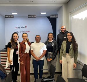 A imagem mostra seis pessoas em pé, na recepção da clínica Grupo Pulsa. Todas vestem roupas em tons neutros e terrosos. Ao fundo, na parede, aparecem logotipos e a frase "aqui a vida pulsa", que compõe a identidade visual do ambiente.
