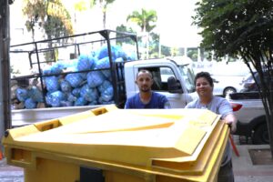 A imagem mostra um caminhão branco de carroceria aberta com grades estacionado junto ao meio-fio. A caçamba está cheia de sacos plásticos azuis empilhados, contendo materiais recicláveis, entre eles os papéis do edital de eliminação. 