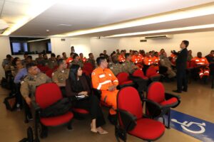  A imagem mostra o auditório do TJMMG com uma plateia de policiais e bombeiros militares sentados em fileiras de poltronas vermelhas. Todos vestem uniformes militares. Ao centro do auditório está o desembargador Fernando Antônio Nogueira Galvão da Rocha, em pé. Ele usa um terno preto e sapato social preto. O auditório tem teto e paredes brancas.