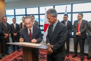 A imagem mostra, em primeiro plano, o desembargador Marco Antônio Borges, usando terno escuro, camisa branca e gravata listrada em vermelho e preto, assinando um documento sobre uma tribuna de madeira, com uma caneta na mão. Ao seu lado está o desembargador Vicente de Oliveira Silva, vestindo terno escuro, camisa cinza e gravata escura. Ao fundo, aparecem bandeiras, além de autoridades, entre homens e mulheres. O primeiro é o desembargador Jãmes Ferreira Santos, vestindo terno preto e gravata escura.